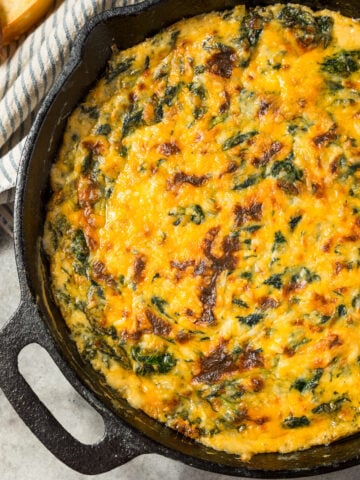 Creamy baked Spinach Maria Dip in a cast iron skillet topped with melted cheddar and mozzarella cheese, ready to serve with bread and chips.