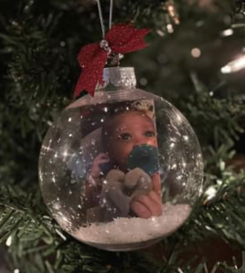 DIY clear snow globe photo ornament with a baby photo inside, faux snow at the bottom, and a red glitter bow on top hanging on a Christmas tree
