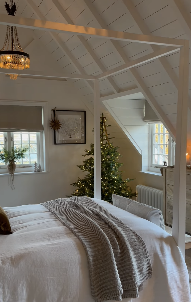 cozy farmhouse christmas bedroom with warm lighting, neutral bedding, and lit christmas tree by window