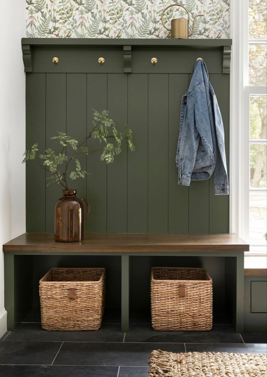 Mudroom with dark green shiplap, built-in wood bench, wicker storage baskets, brass hooks, and floral wallpaper above.