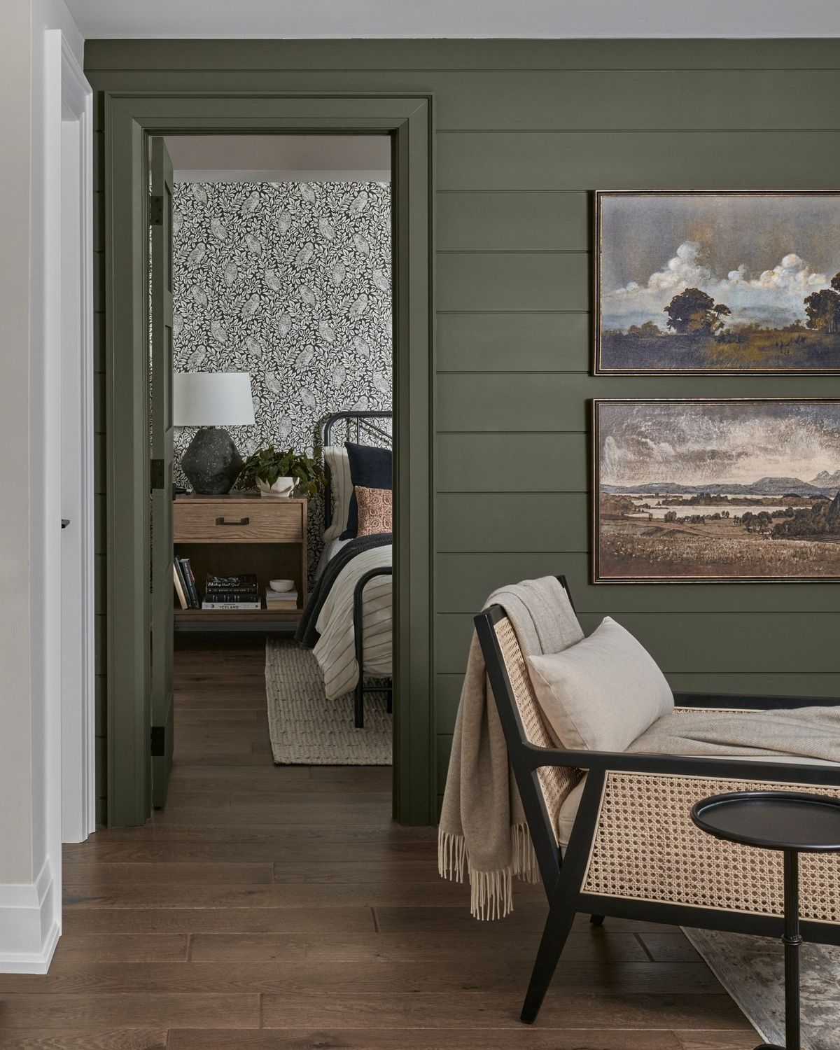 Dark green shiplap accent wall with doorway opening into patterned bedroom, wood flooring, and neutral woven lounge chair.