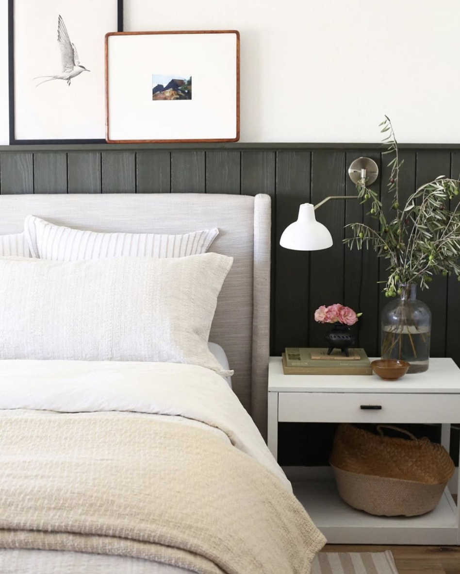 Bedroom with dark green vertical shiplap half wall, white bedding, brass sconce lighting, and minimalist artwork.