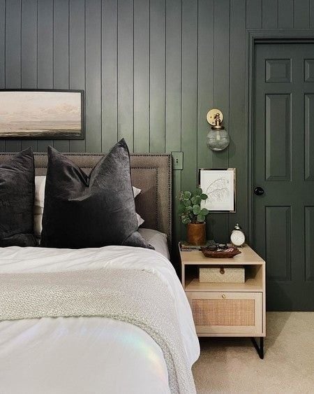 Cozy bedroom with dark green vertical shiplap wall, upholstered headboard, black velvet pillows, and rattan nightstand.
