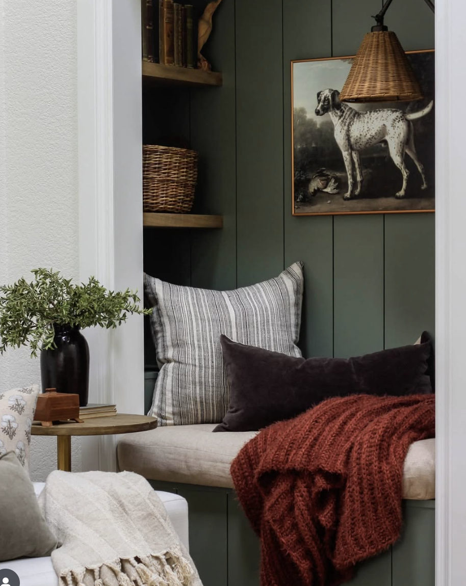 Cozy reading nook with dark green vertical shiplap, built-in shelves, striped pillows, wicker pendant light, and vintage dog artwork.