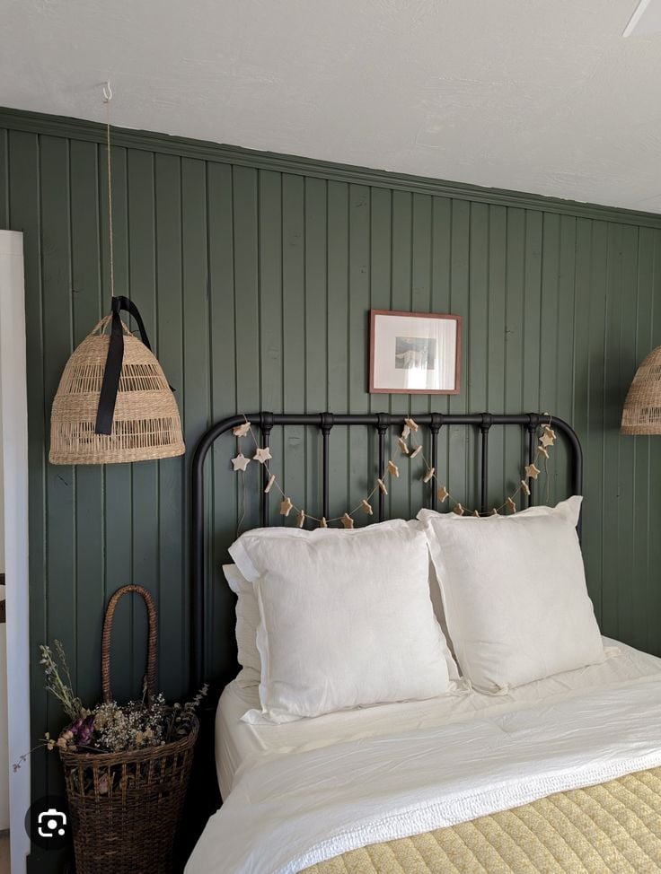 Cottage bedroom with dark green vertical shiplap walls, black metal bed frame, white bedding, and wicker pendant lights.