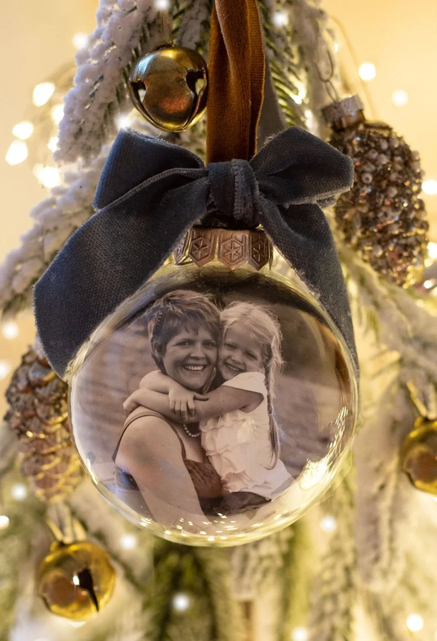 DIY clear glass globe photo ornament with black and white family photo inside, tied with a navy velvet ribbon and hanging on a Christmas tree