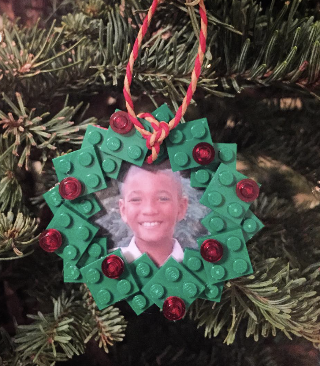 DIY LEGO wreath photo ornament made with green LEGO bricks, red accent pieces, and a child's photo framed in the center hanging on a Christmas tree