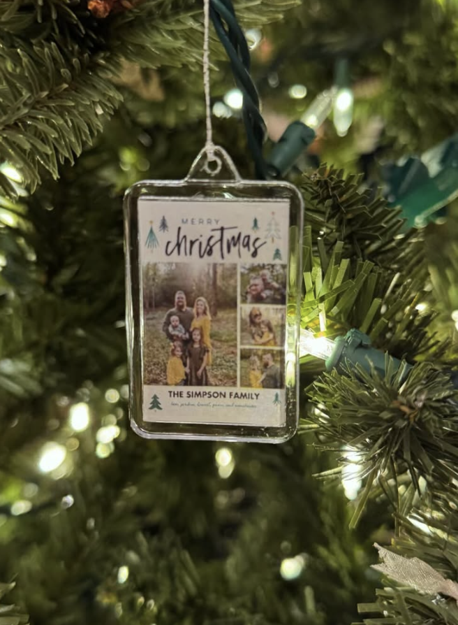 DIY glass frame photo ornament hanging on a Christmas tree, featuring a family holiday photo card with 'Merry Christmas' and greenery accents