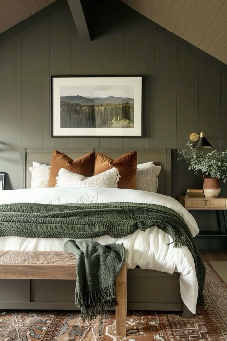 Bedroom with dark green vertical shiplap wall, rust-colored throw pillows, framed landscape art, and modern brass sconce lighting.