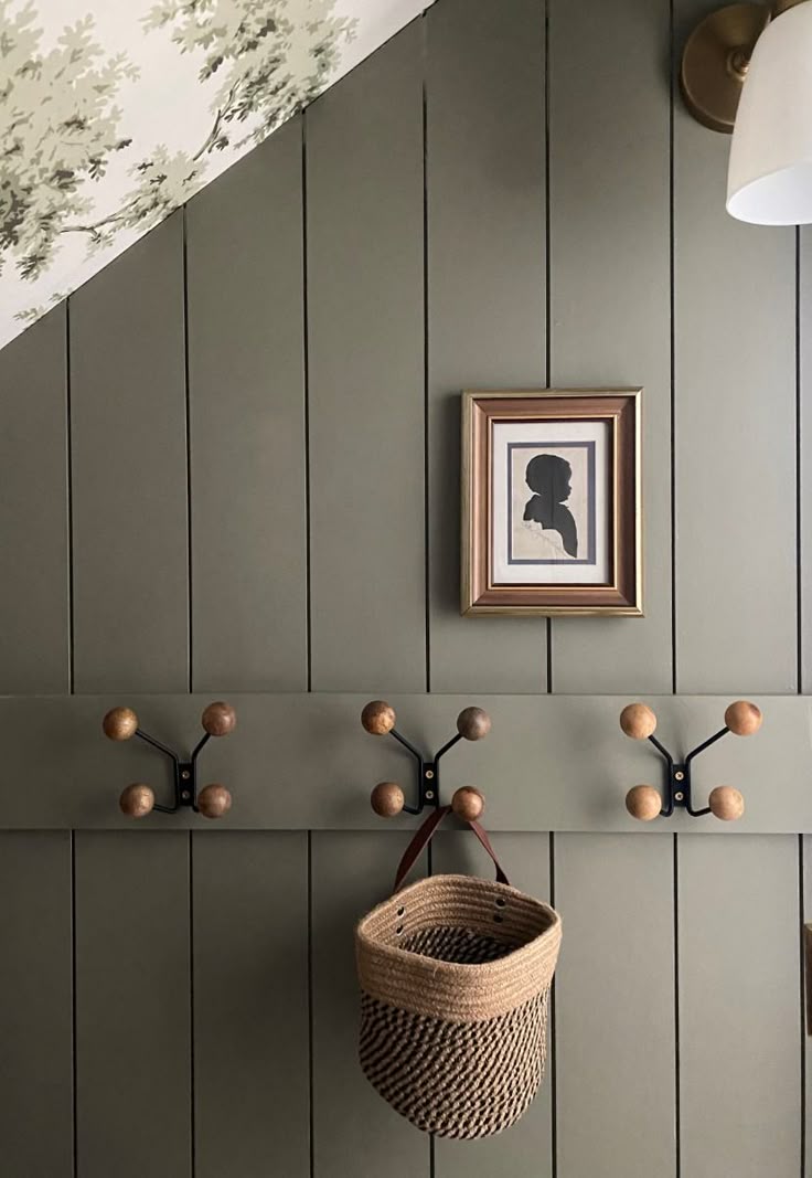 Green vertical shiplap wall with wood peg rail, woven basket, brass sconce, and small framed silhouette artwork.