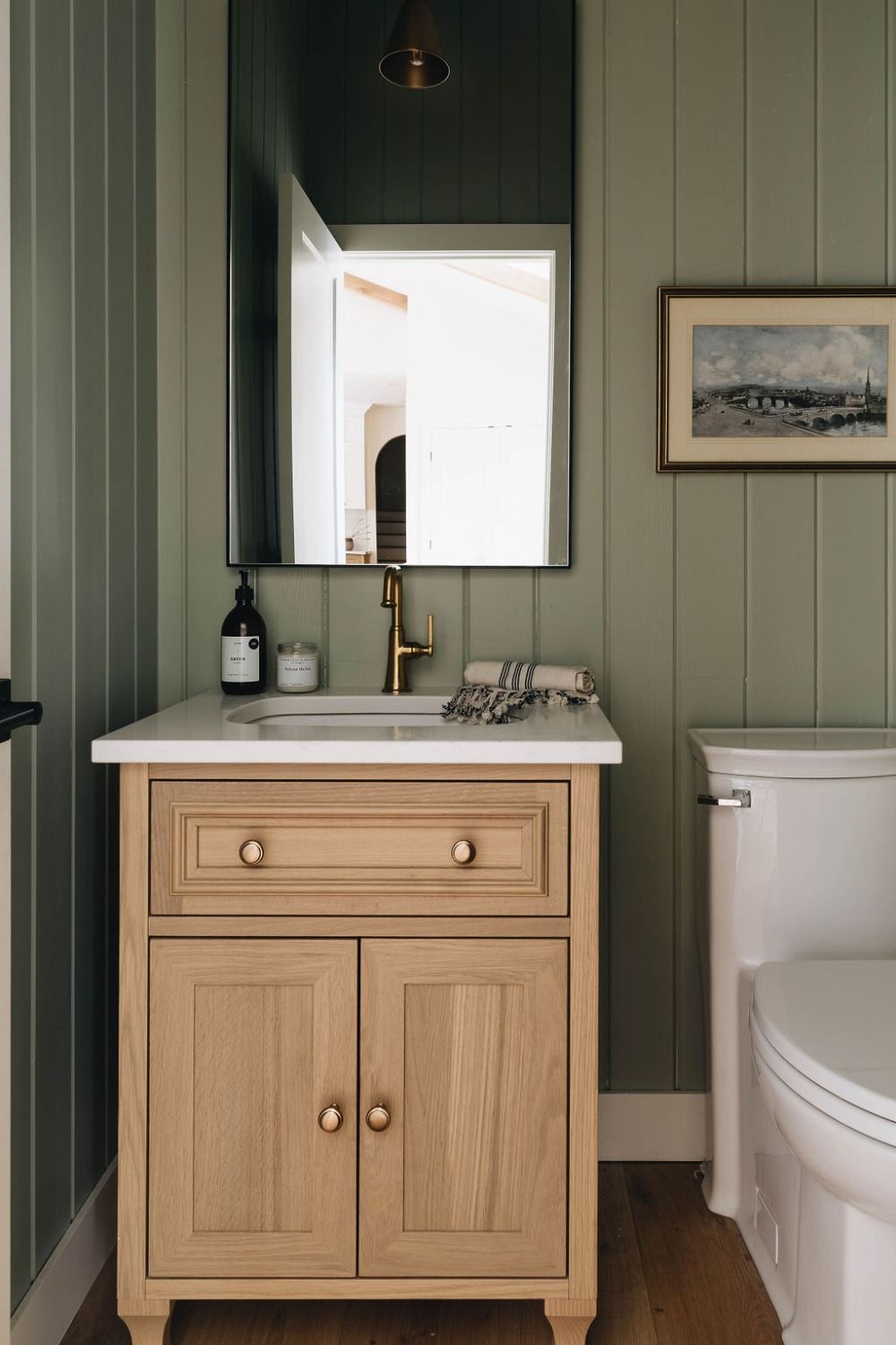 Bathroom with light green vertical shiplap walls, natural wood vanity, brass faucet, and framed art above the toilet.