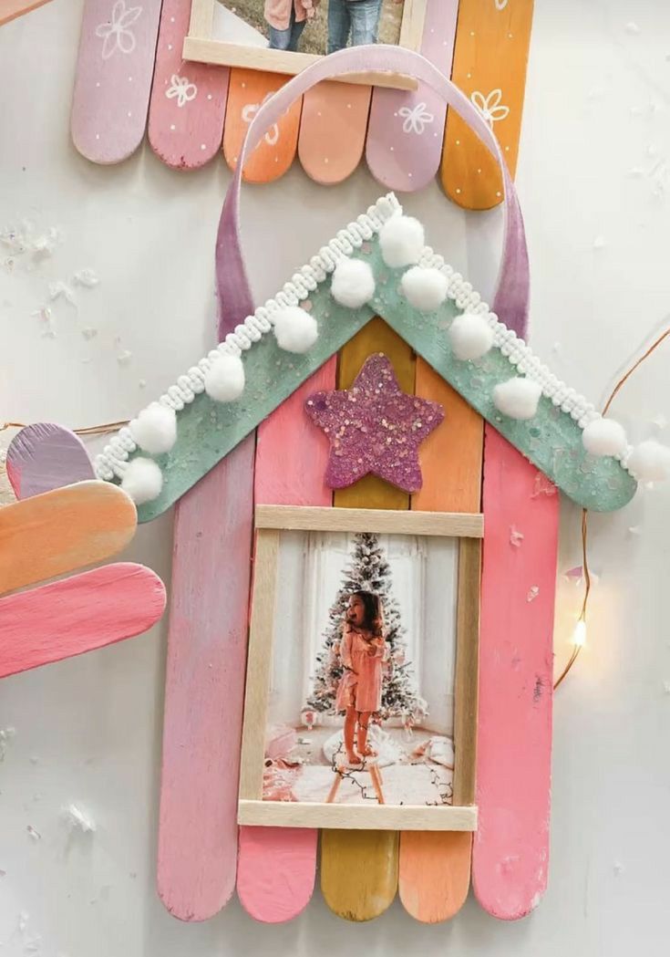 DIY pastel popsicle stick photo ornament shaped like a house, decorated with pom-poms, glitter star, and a Christmas photo of a little girl by the tree