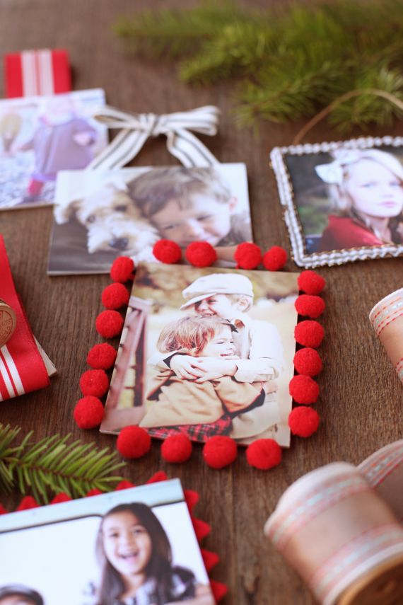 DIY photo ornaments decorated with red pom-pom trim, ribbon bows, and family photos laid out on a wooden table with greenery and craft supplies