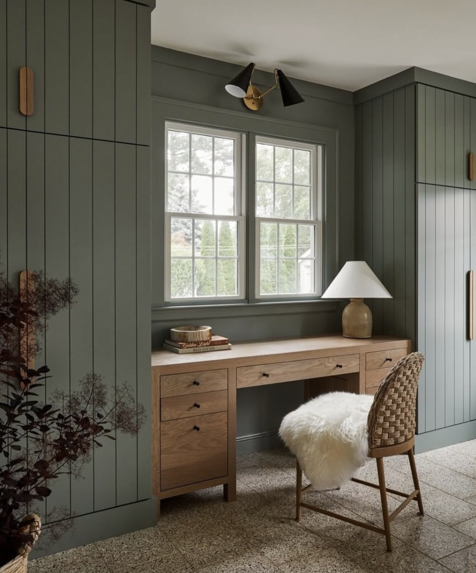 Living room with sage green vertical shiplap walls, built-in wood desk, woven chair with fur throw, and black and brass wall sconce.