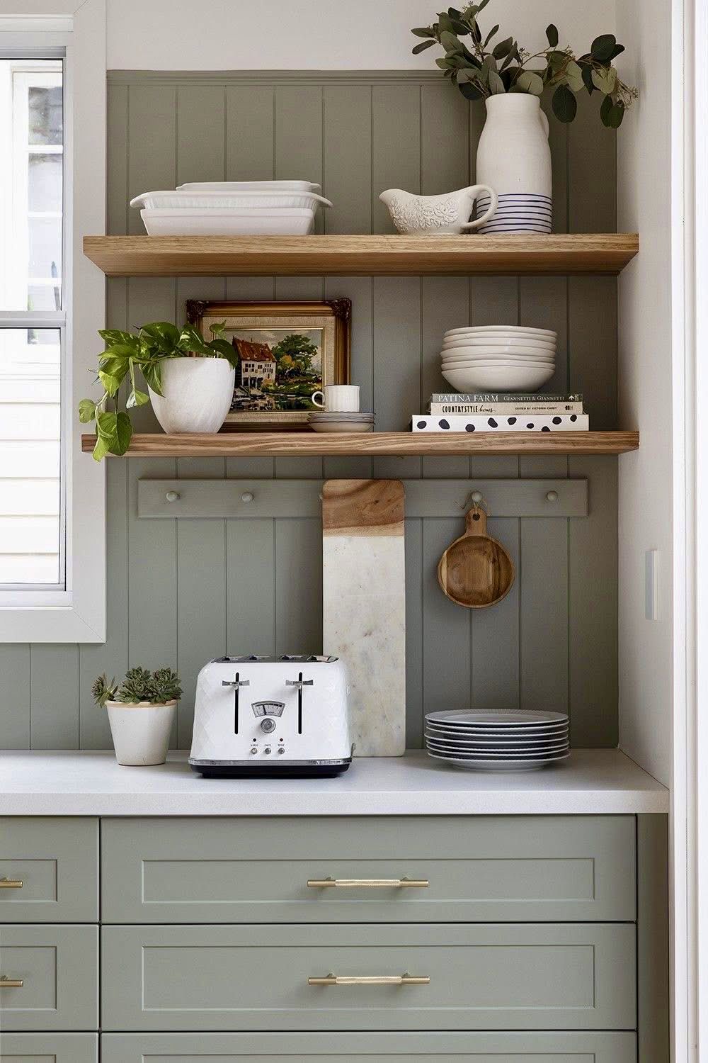 Kitchen with sage green vertical shiplap backsplash, wood open shelving, white dishes, and sage cabinetry with brass hardware.