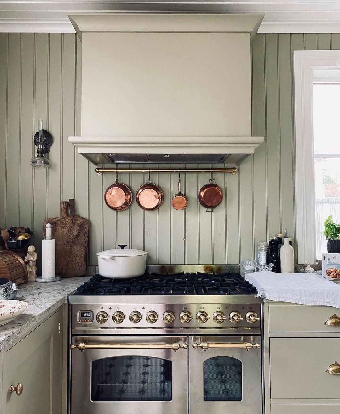 Kitchen with sage green vertical shiplap walls, brass range hood rail displaying copper pots, stainless steel range, and stone countertops.