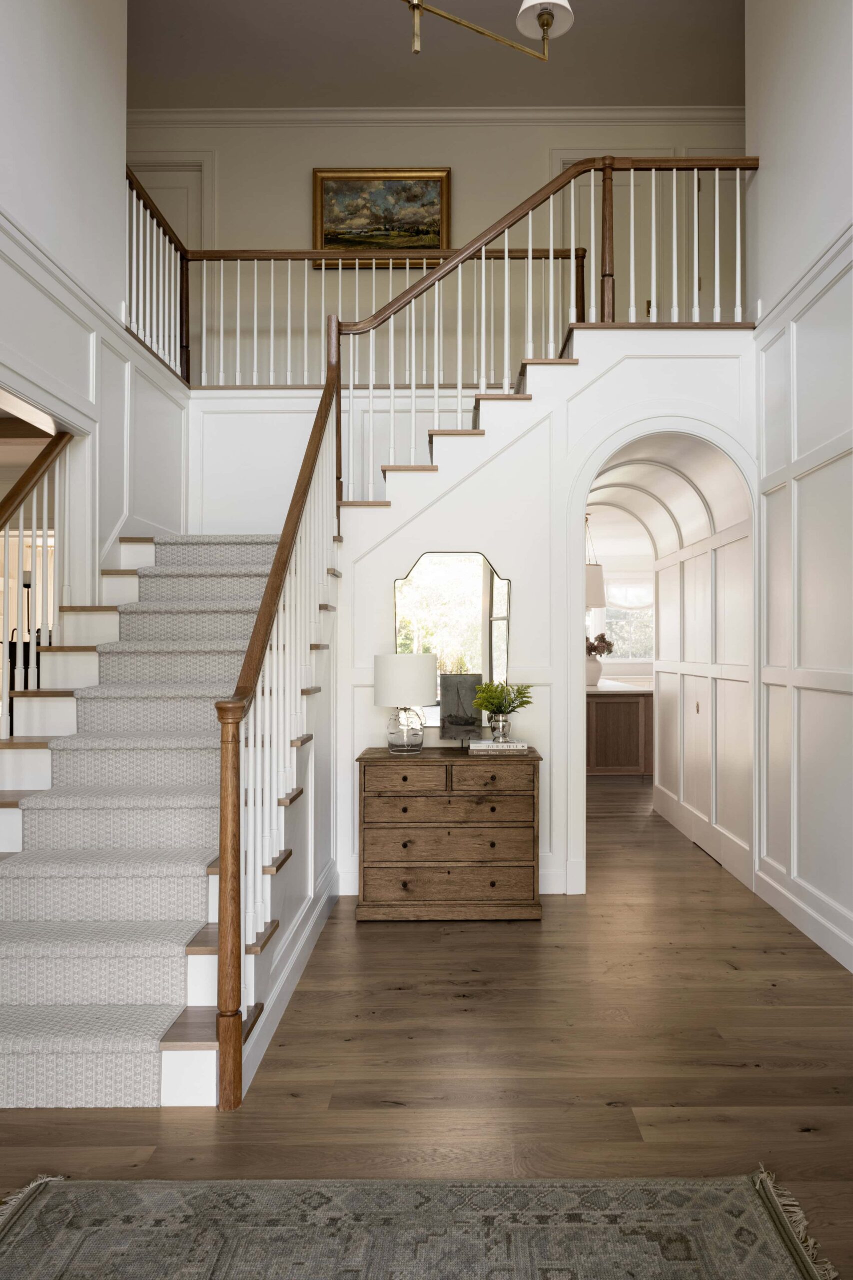 Studio McGee entryway with arched hallway, wood dresser, mirror, white panelled walls, and gray stair runner creating a bright classic foyer.
