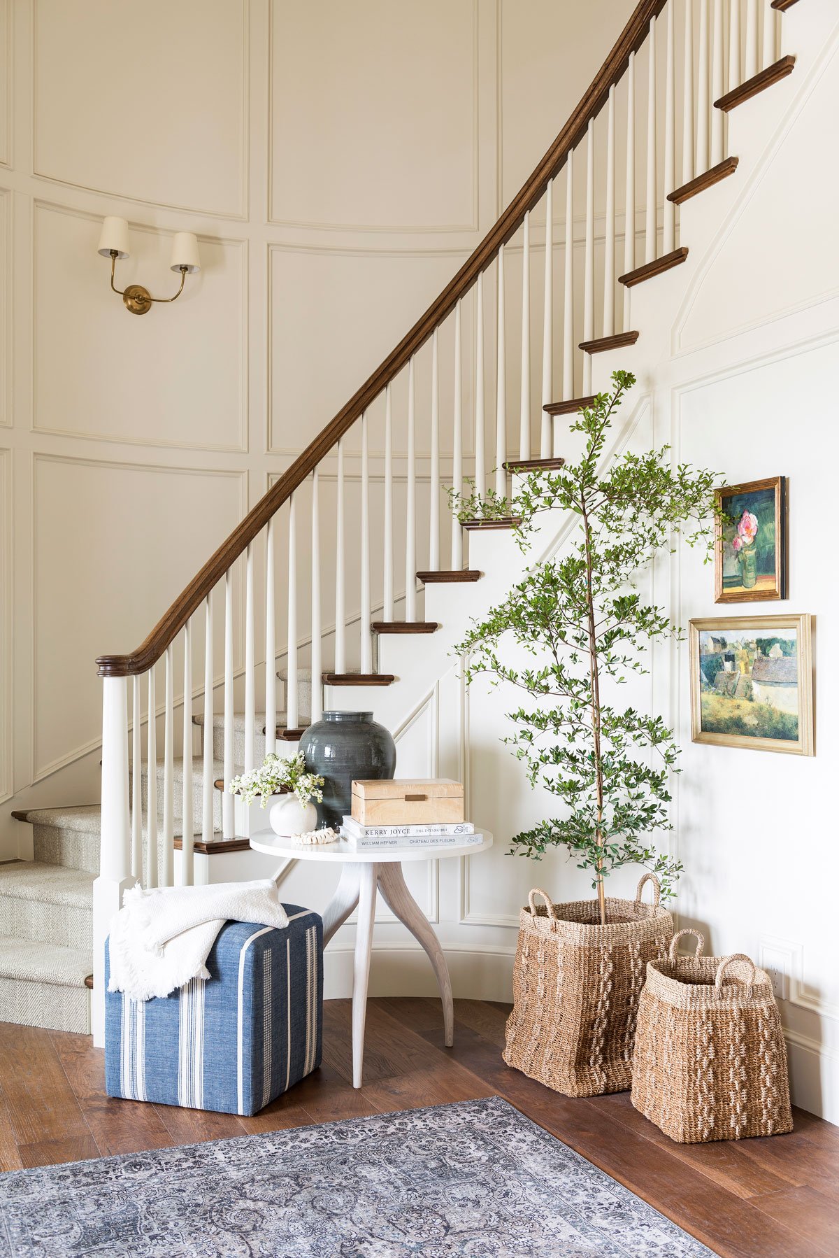 Studio McGee foyer with curved staircase, round entry table, woven baskets, blue ottoman, and layered artwork creating a warm, inviting entryway.