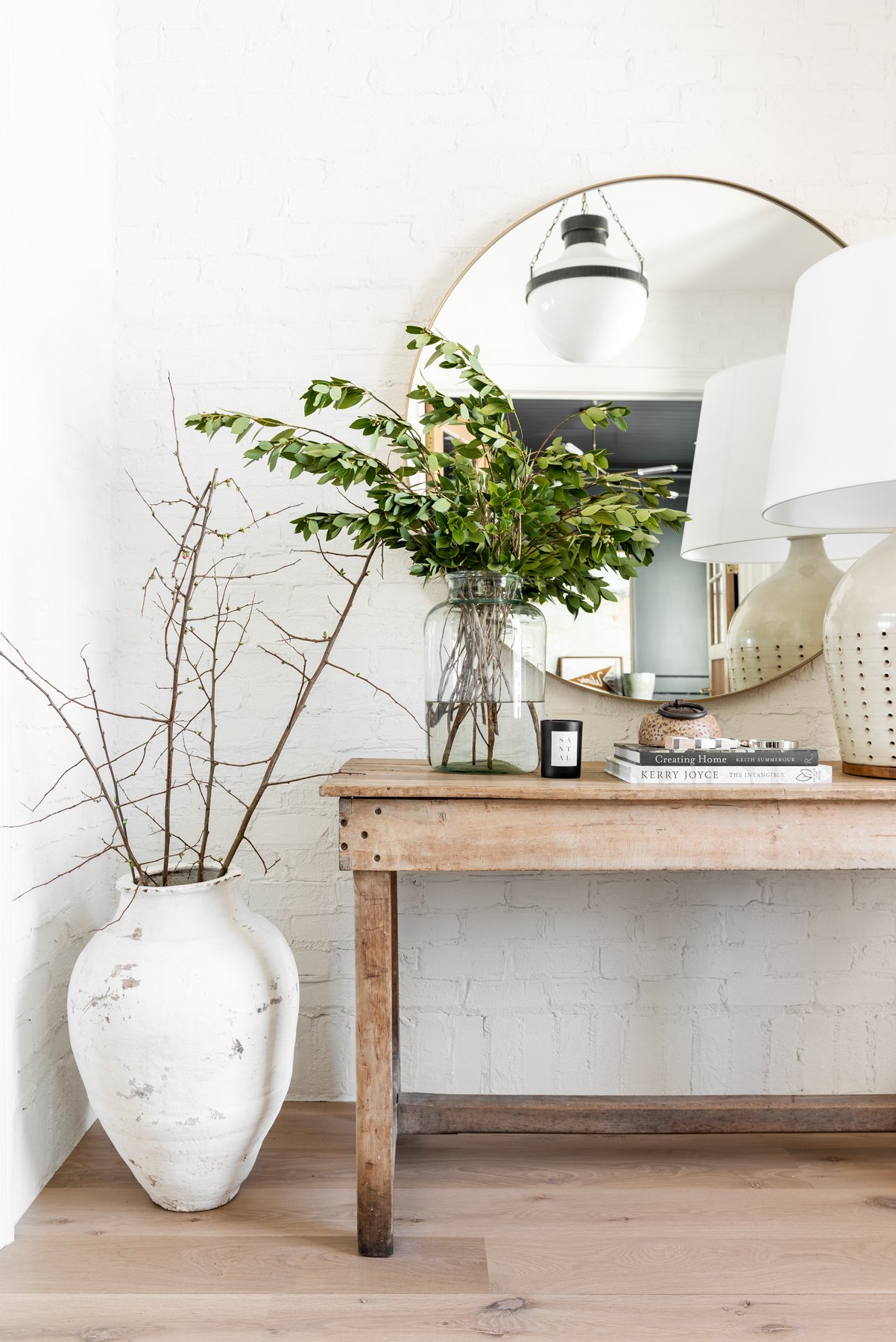 Studio McGee entryway with white brick wall, rustic wood console, glass vase with greenery, round mirror, and large clay pot for a natural, minimal look.