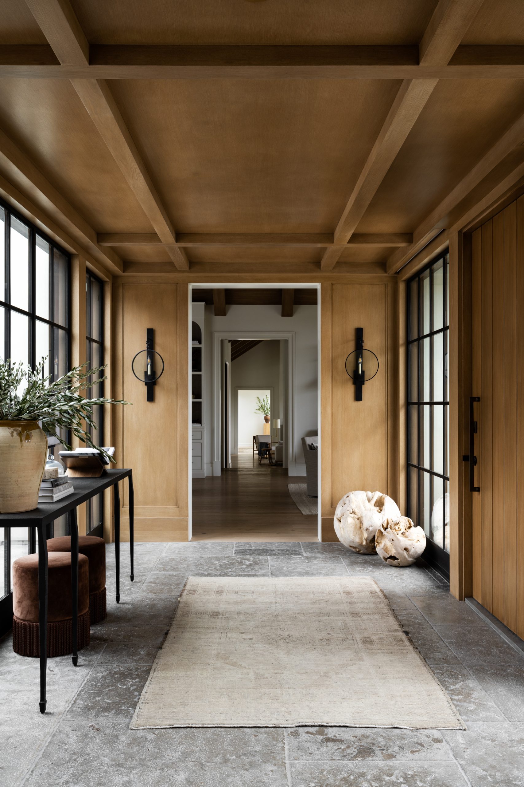 Modern Studio McGee entryway with wood paneling, black console table, greenery, vintage rug, and large black-framed windows creating a warm sophisticated foyer.