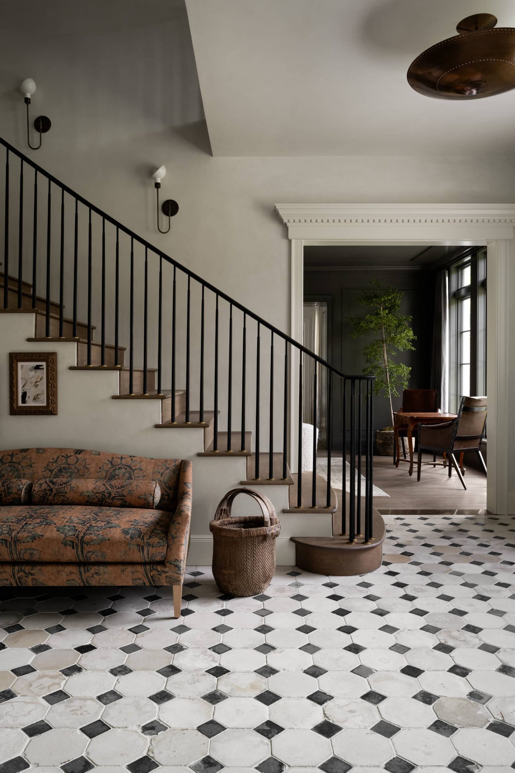 Studio McGee entryway with black and white patterned tile floor, curved staircase with iron railing, vintage floral sofa, and woven basket for a European-inspired design.