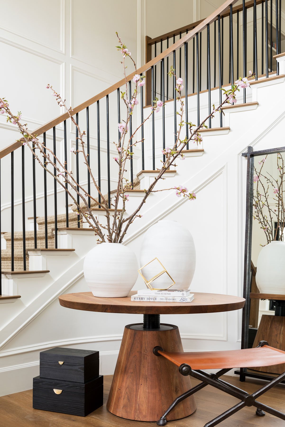 Studio McGee grand entryway featuring round wood table with white vases and flowering branches, black stair railing, and warm modern farmhouse design.