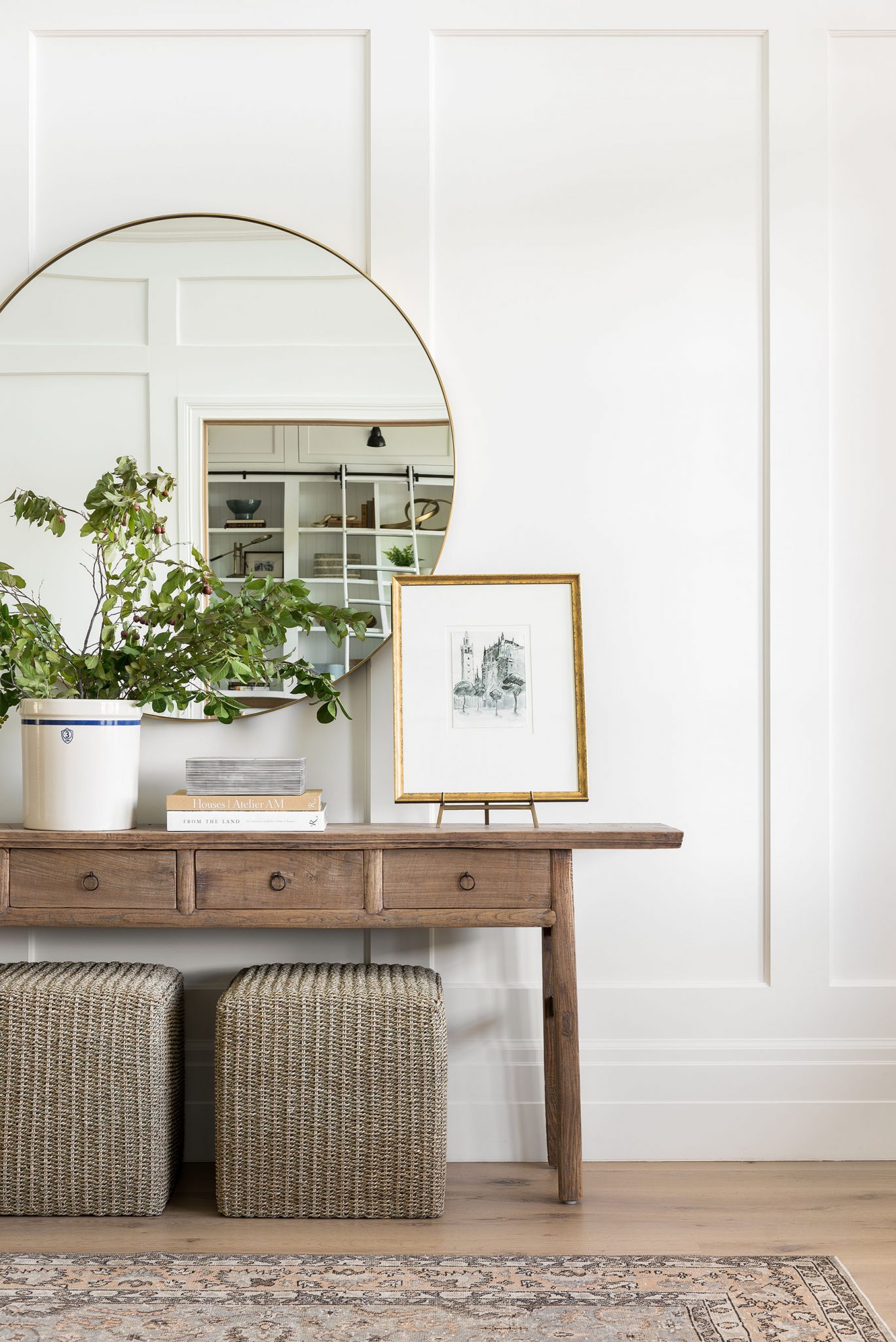 Studio McGee entryway featuring rustic wood console table, large round mirror, woven stools, and greenery on white paneled wall for a bright, welcoming look.