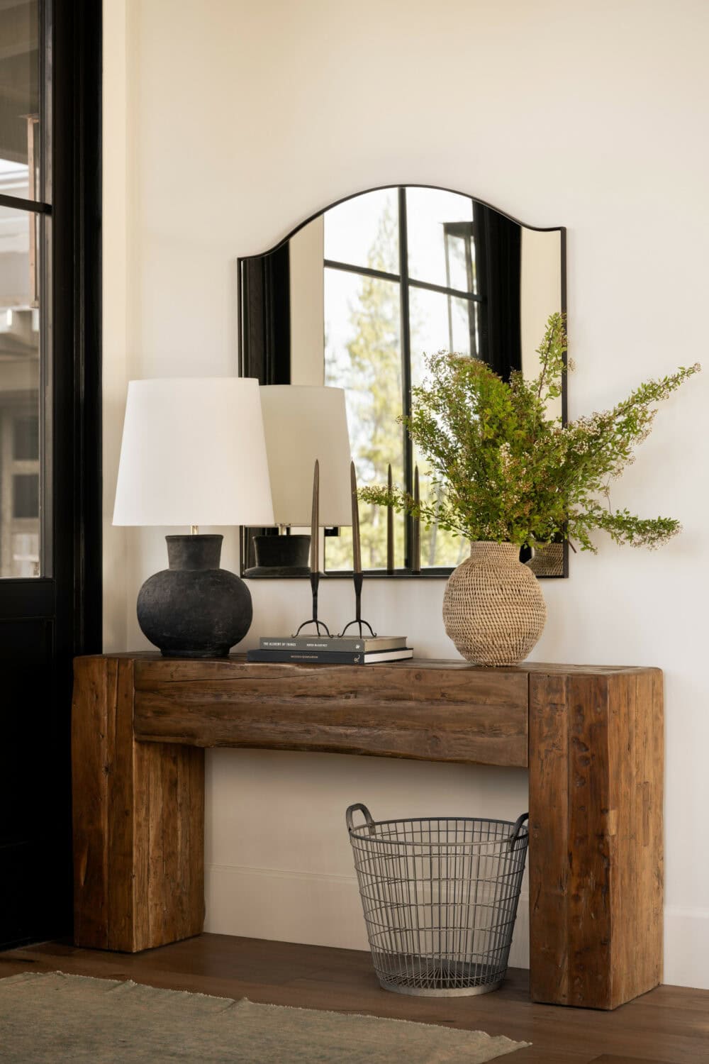 Studio McGee entryway with rustic wood console, black lamp, woven vase with greenery, and arched mirror creating a warm, minimalist foyer.