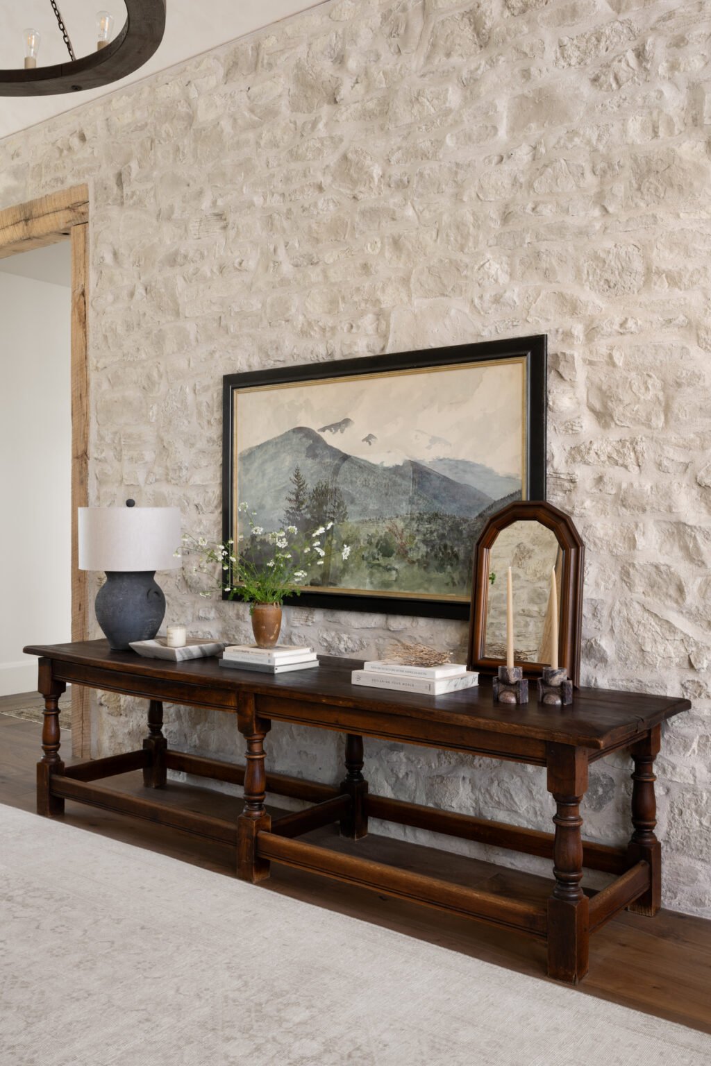 Studio McGee entryway with natural stone wall, dark wood console table, vintage mirror, framed landscape art, and vase of wildflowers for a timeless rustic look.