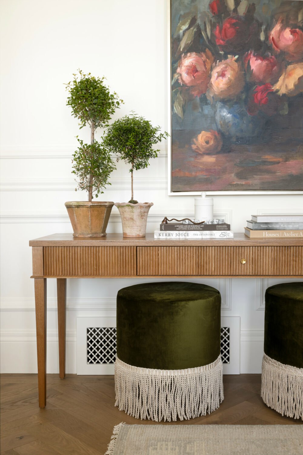 Studio McGee entryway featuring fluted wood console table, topiary plants, stacked design books, and green velvet stools with fringe under large floral artwork.