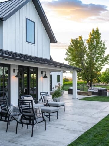 Modern farmhouse backyard with white board-and-batten siding, black trim, and a spacious concrete patio featuring outdoor lounge chairs, greenery, and cozy seating areas at sunset.