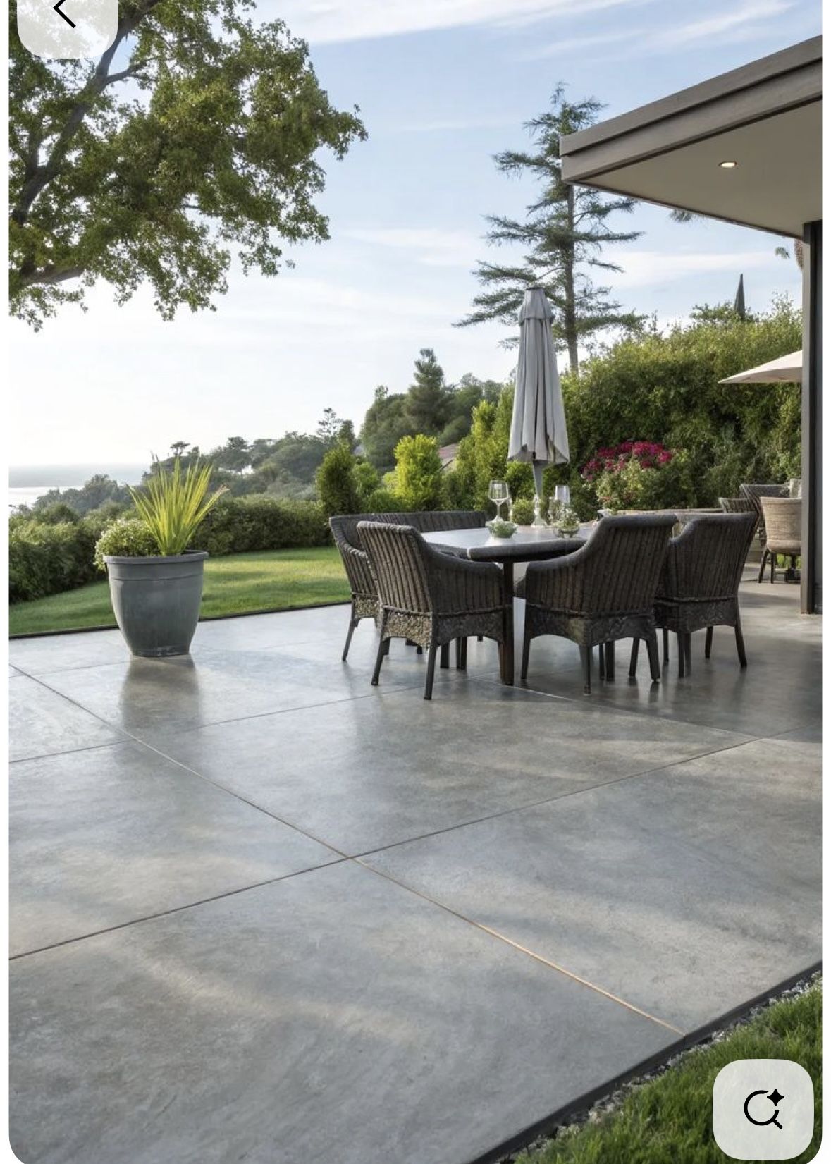 Modern outdoor patio with large decorative concrete slabs, wicker dining chairs, a round table, and surrounding greenery overlooking a scenic landscape.