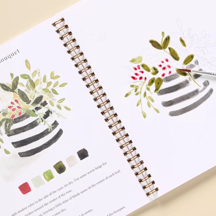 Open Emily Lex watercolor workbook showing floral painting steps and color swatches with a paintbrush in progress.