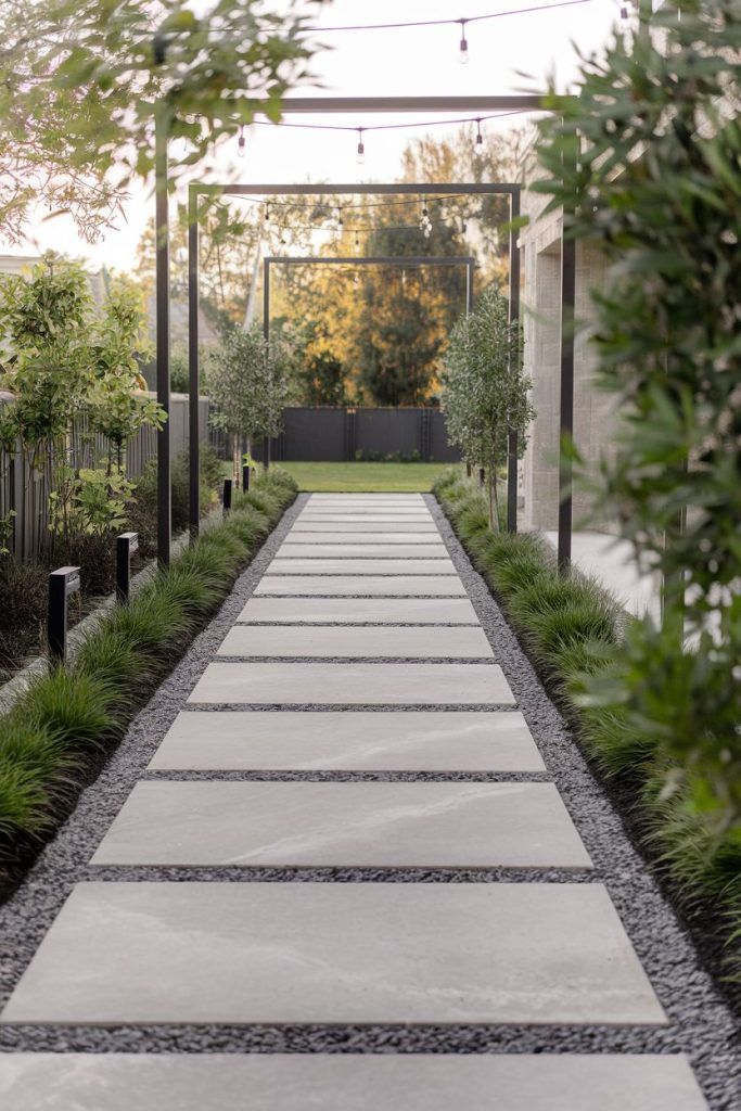 Long modern garden pathway made of large concrete stepping stones set in gravel, lined with lush greenery and overhead string lights for a clean, structured outdoor look.