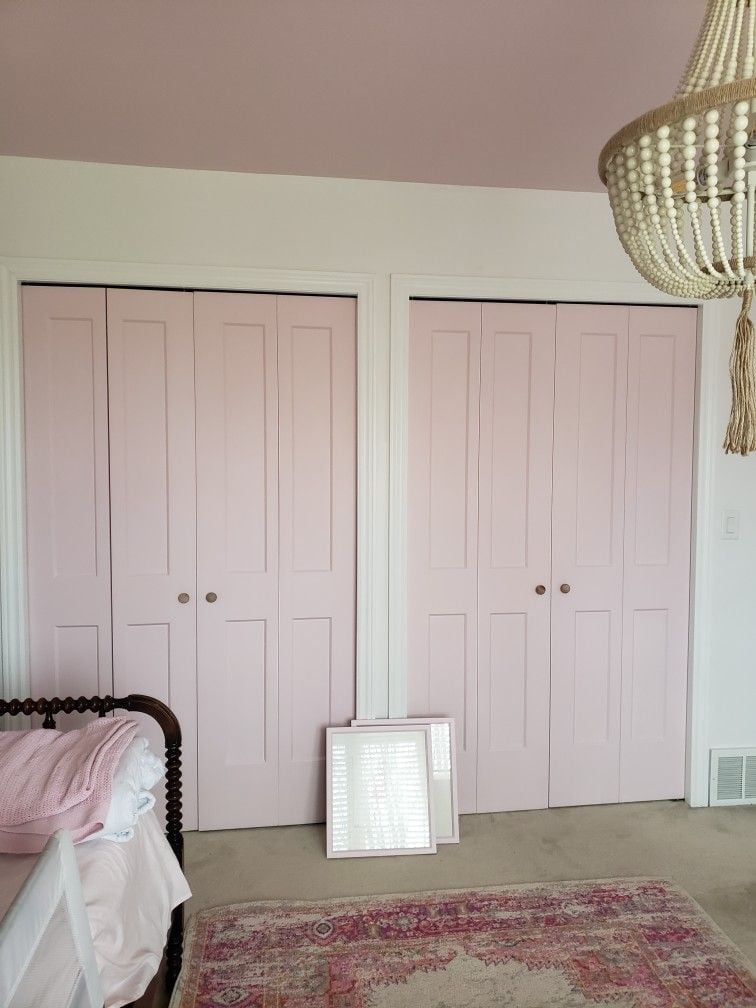 Light pink bifold closet doors with brass knobs in a soft, feminine bedroom featuring a beaded chandelier, vintage rug, and warm neutral decor.