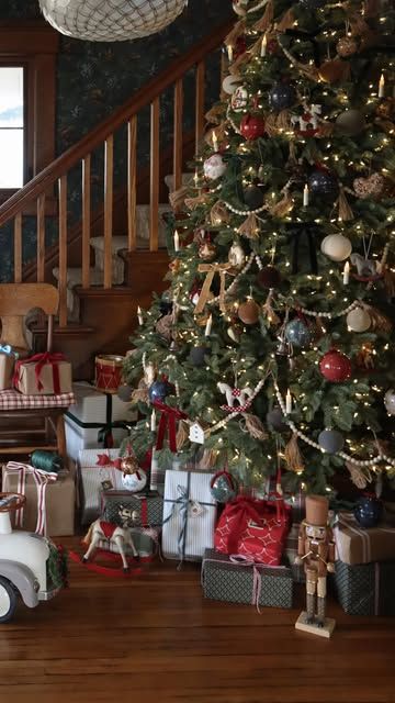 Classic Ralph Lauren-style Christmas tree decorated with plaid ornaments, gold garland, and warm white lights, surrounded by wrapped gifts, a nutcracker, and vintage toys on a hardwood floor beside a staircase.