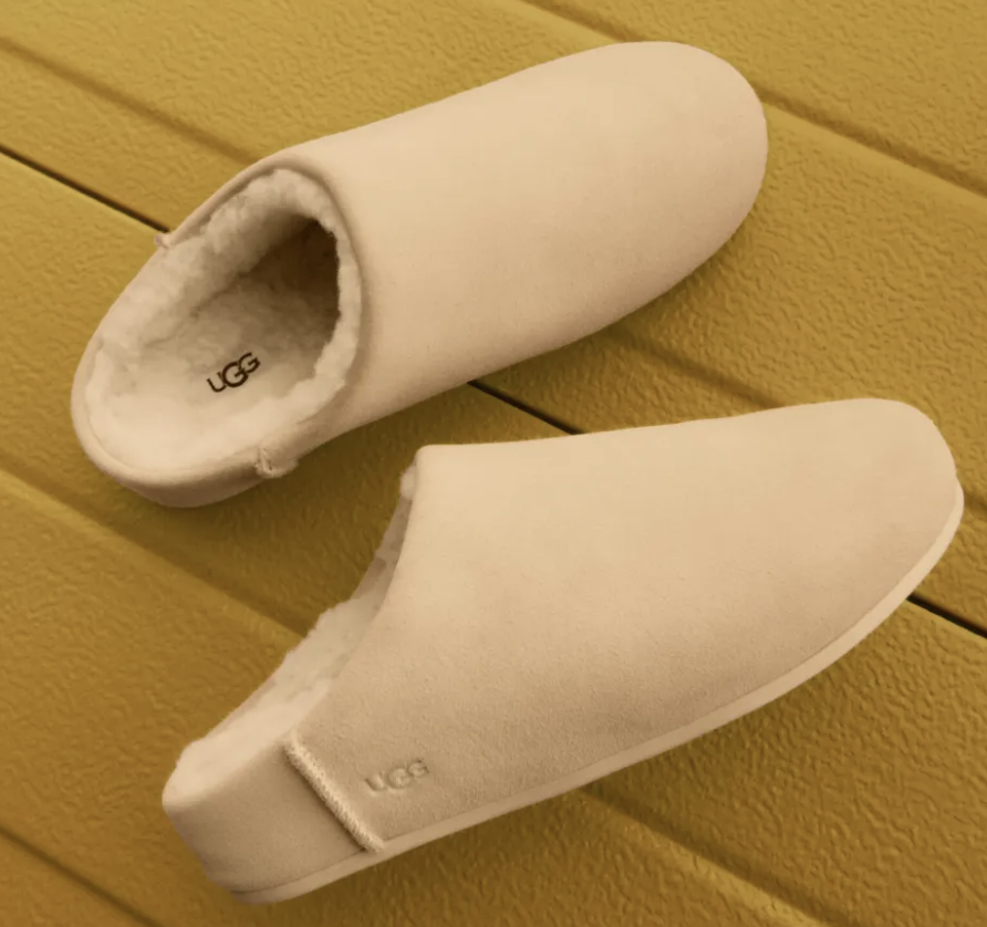 UGG Elea slip-on shearling slippers in cream with cozy lining on a warm yellow background.