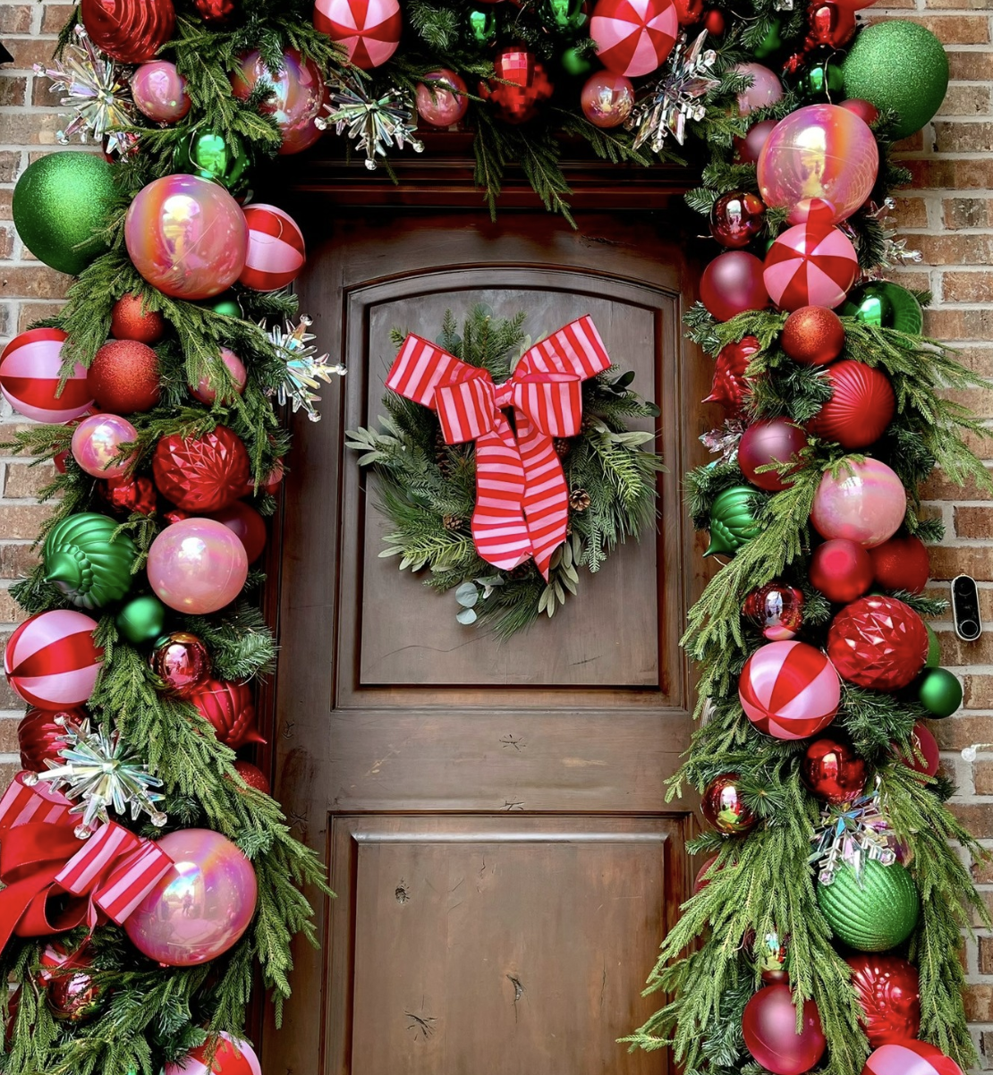 Wicked-inspired Christmas front door decorated with pink, green, and red ornaments, cedar garland, and a striped red bow wreath.