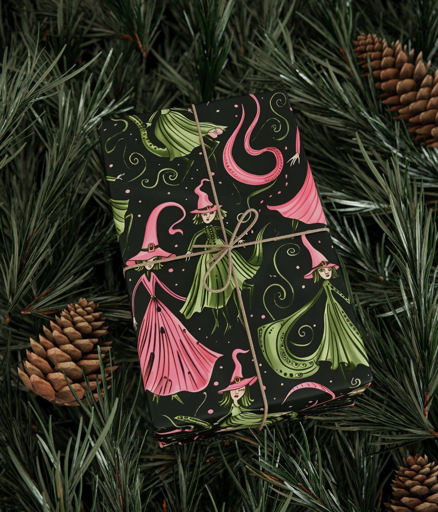 Wicked-themed Christmas gift wrapped in black paper with pink and green illustrated witches, tied with twine and surrounded by pine greenery.