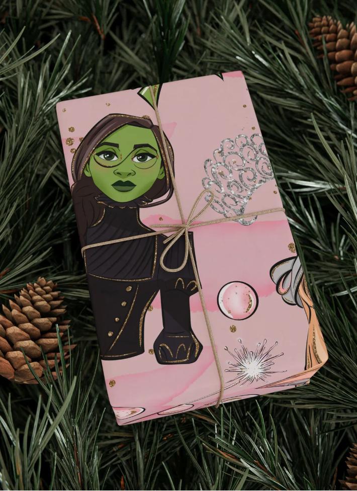 Pink Wicked-themed Christmas gift wrap featuring an illustration of Elphaba with green skin, tied with twine and surrounded by pine branches.