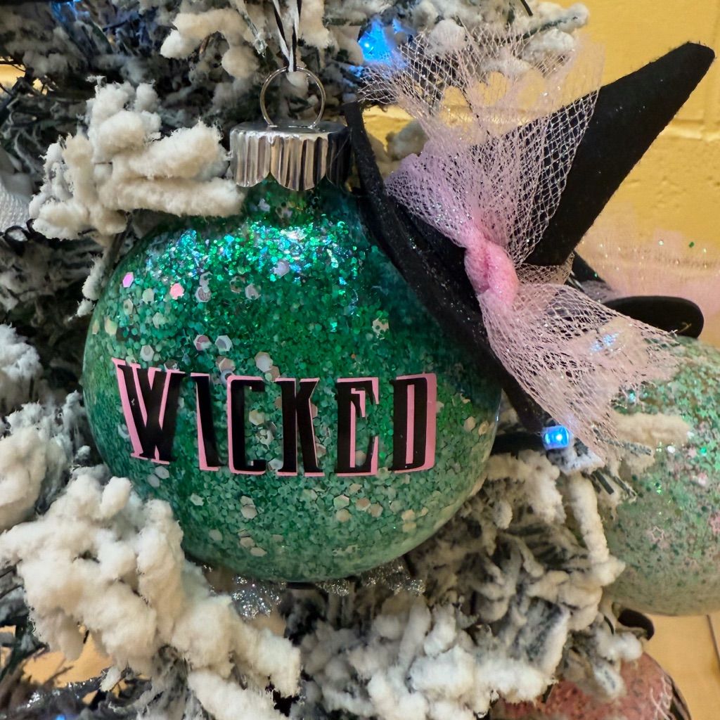 Green glitter Wicked Christmas ornament with pink lettering and a small witch hat, hanging on a snow-covered Christmas tree.
