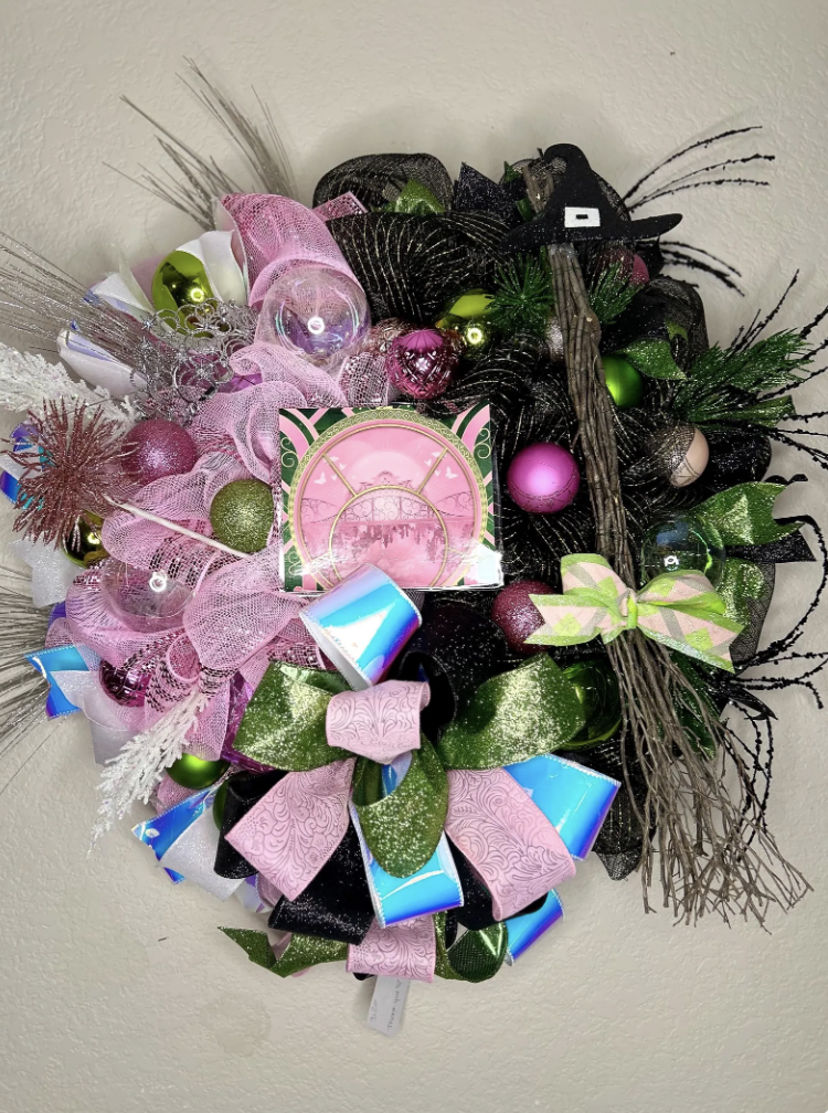 Wicked Christmas wreath featuring pink, green, and black glitter ribbon, colorful ornaments, and a small witch broom accent.