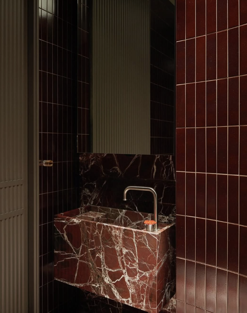 A modern powder room featuring a deep red marble sink with bold white veining, glossy red vertical tile walls, and a sleek brushed metal faucet.