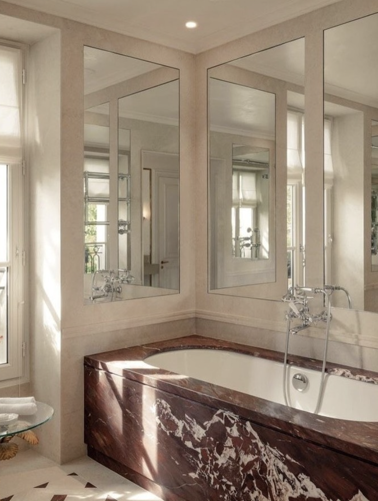 A refined bathroom featuring a red marble bathtub surround with bold white veining, large framed mirrors, neutral plaster walls, and polished chrome fixtures.