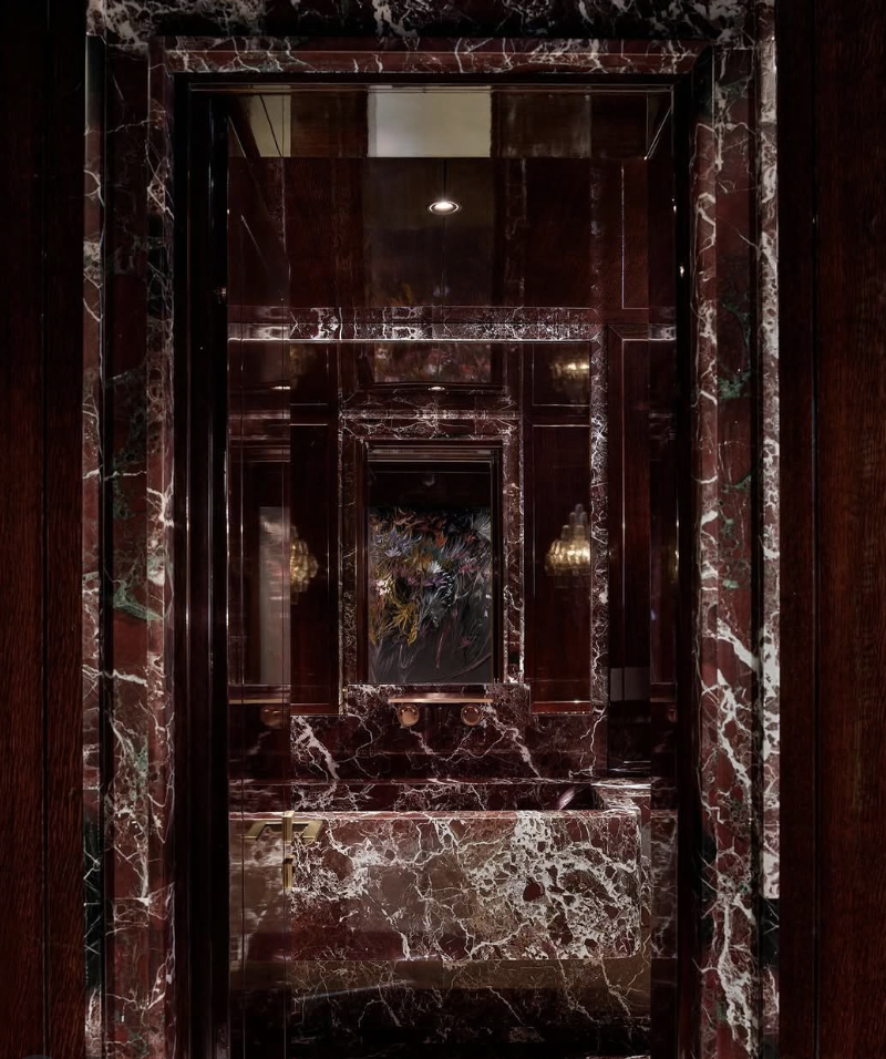 A dramatic luxury bathroom fully clad in dark red marble with bold white veining, featuring a marble-wrapped vanity, rich wood accents, brass hardware, and mirrored reflections.