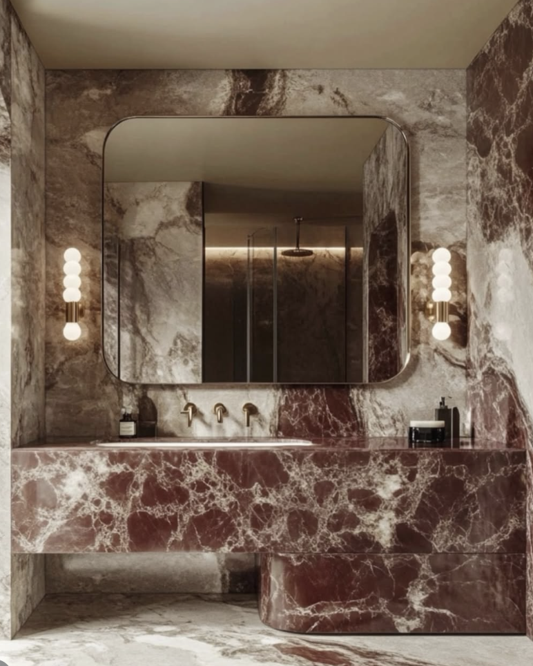 A high-end bathroom featuring a bold red marble vanity with white veining, full marble wall cladding in taupe and red tones, brass fixtures, globe sconces, and a rounded rectangular mirror.