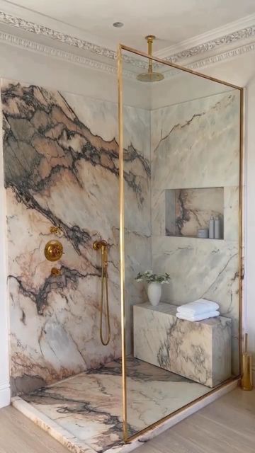 A high-end marble shower featuring soft cream stone with dramatic red and charcoal veining, brass shower fixtures, a built-in shower bench, and a brass-framed glass enclosure.
