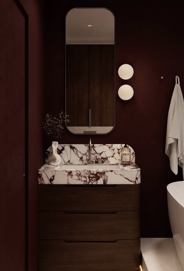A contemporary bathroom featuring a rounded wood vanity topped with red and white marble, set against deep burgundy walls with a tall modern mirror and globe sconces.