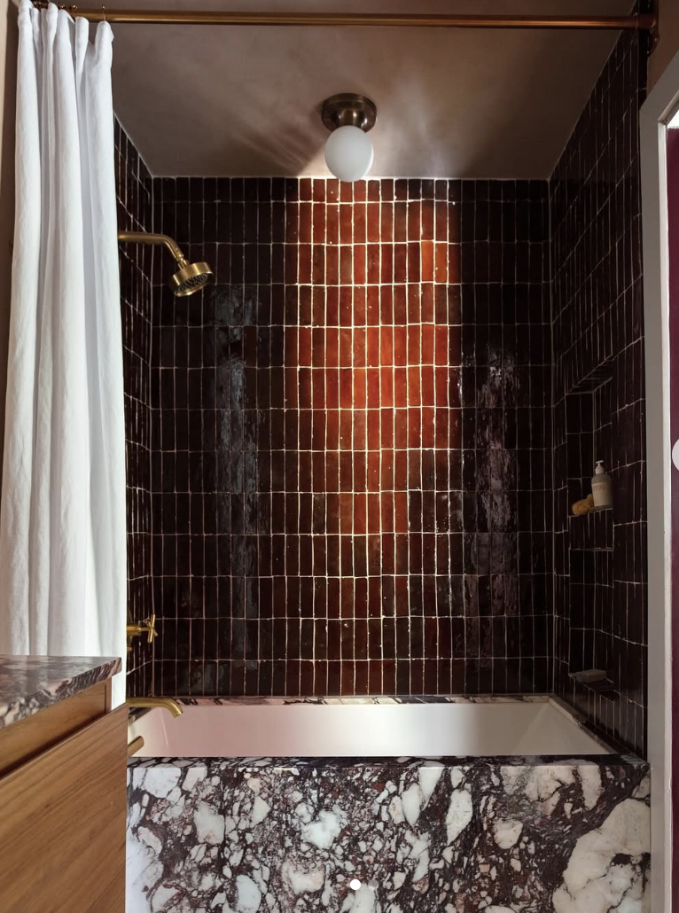 A dramatic bathroom featuring glossy deep red shower tile, a red-and-white marble tub surround, brass fixtures, and a simple globe ceiling light.