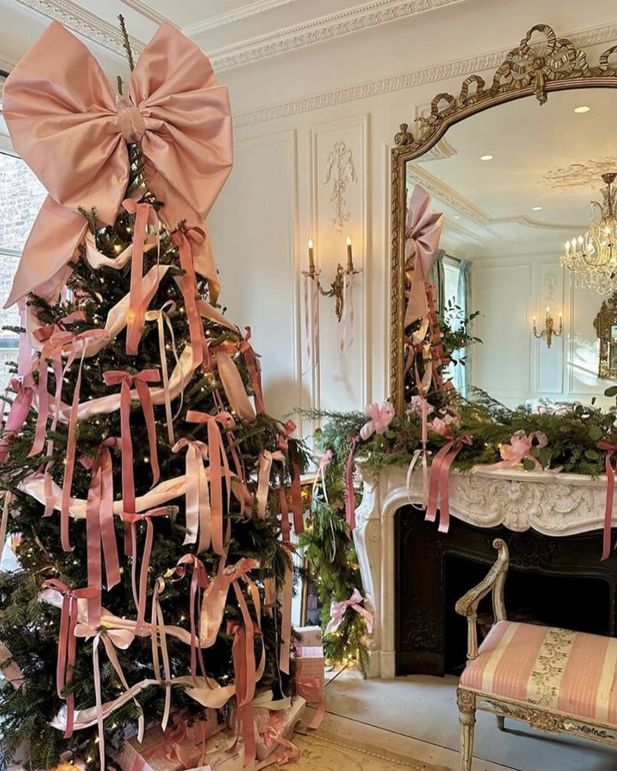 Elegant Christmas tree decorated with an oversized pink bow topper, cascading satin ribbons, and a matching pink ribbon garland on a vintage-style mantel.